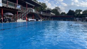 Blick vom Kopf des großen 50m Beckens auf das Becken. Links am Beckenrand sind einige Aussteller-Stände zu sehen. Rechts im Bild, im Wasser, sieht Luna schwimmen. Luna ist zu dem Zeitpunkt der einzige Hund im Wasser.