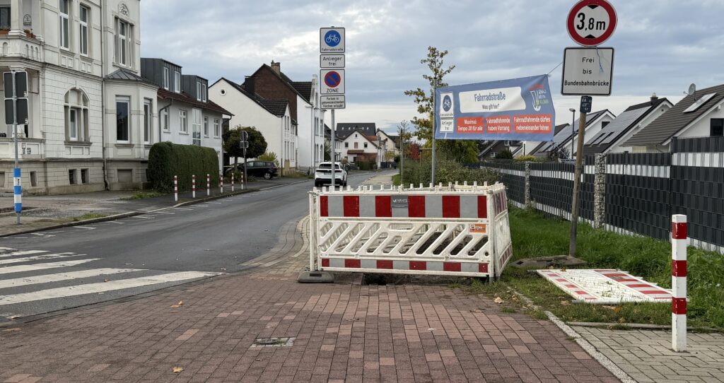 abgesperrte Baustelle; die Absperrung blockiert fast den gesamten Bürgersteig. Weiterkommen mit Kinderwagen, Rollstuhl oder Rollator ist nur über die Straße möglich